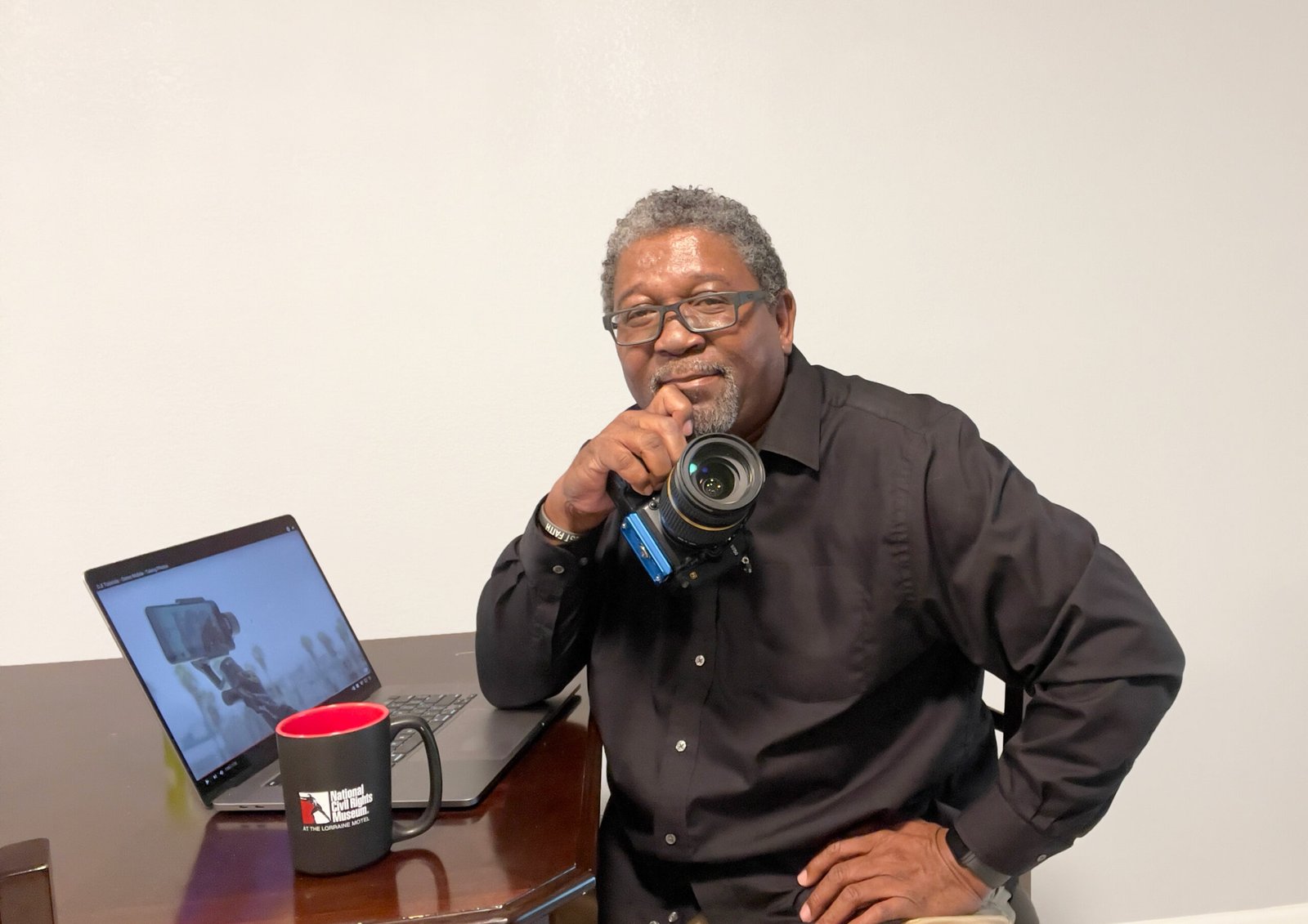 rafael-sitting-at-table-holding-camera Rafael Powell with camera, laptop and a cup of coffee